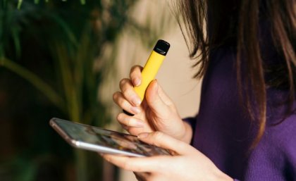 A young woman in profile with long dark hair falling across her face, holding a vape and a mobile phone.